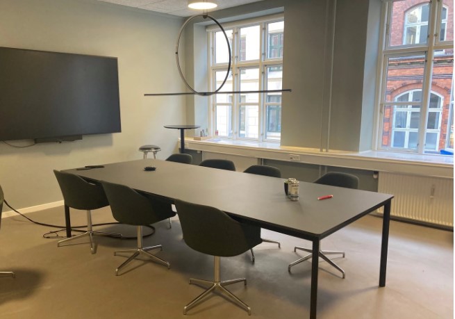 Et funktionelt coworking-space med et stilrent mødelokale, der har et langt mørkt bord og ergonomiske drejestole.